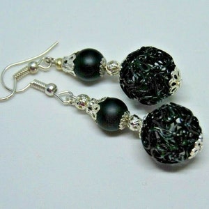 May include: A pair of drop earrings with silver hooks. Each earring has a matte black bead, a cluster of small silver beads, and a dark green filigree ball. The earrings are set against a white backdrop.