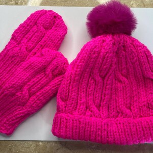 May include: A bright pink knitted hat with a fluffy pom pom and matching mittens. The hat and mittens have a cable knit pattern.