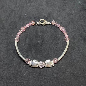 Könnte beinhalten: Ein silbernes Armband mit zwei Fischanhängern und rosa Kristallperlen. Das Armband hat einen silbernen Verschluss. Die Perlen sind klein und gleichmäßig verteilt. Das Armband ist vor einem dunklen Hintergrund platziert.