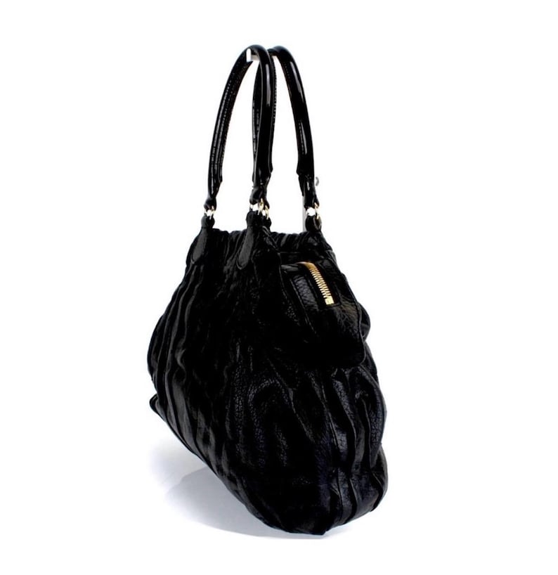 Puede incluir: Bolso de mano de cuero negro con superficie texturizada y cremallera dorada. El bolso tiene dos asas negras y forma redondeada. El bolso est&aacute; sobre un fondo blanco.