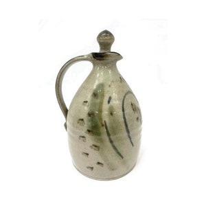 May include: A ceramic pitcher with a lid, featuring a light brown glaze with green and brown abstract designs. The pitcher has a rounded body and a handle.