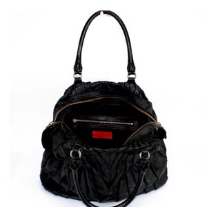Puede incluir: Bolso de mano de cuero negro con dise&ntilde;o fruncido, con cremallera superior y asas dobles. El interior es visible, revelando un peque&ntilde;o bolsillo y una etiqueta roja. El bolso est&aacute; sobre un fondo blanco.