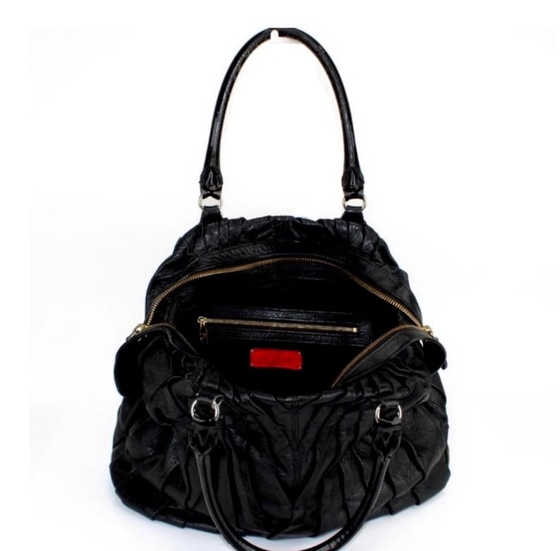 Puede incluir: Bolso de mano de cuero negro con dise&ntilde;o fruncido, con cremallera superior y asas dobles. El interior es visible, revelando un peque&ntilde;o bolsillo y una etiqueta roja. El bolso est&aacute; sobre un fondo blanco.