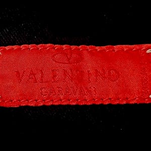 Puede incluir: Primer plano de una etiqueta de tela roja con el texto en relieve "VALENTINO GARAVANI" en may&uacute;sculas. La etiqueta est&aacute; cosida a una tela negra. El logotipo de Valentino es visible en la parte superior de la etiqueta.