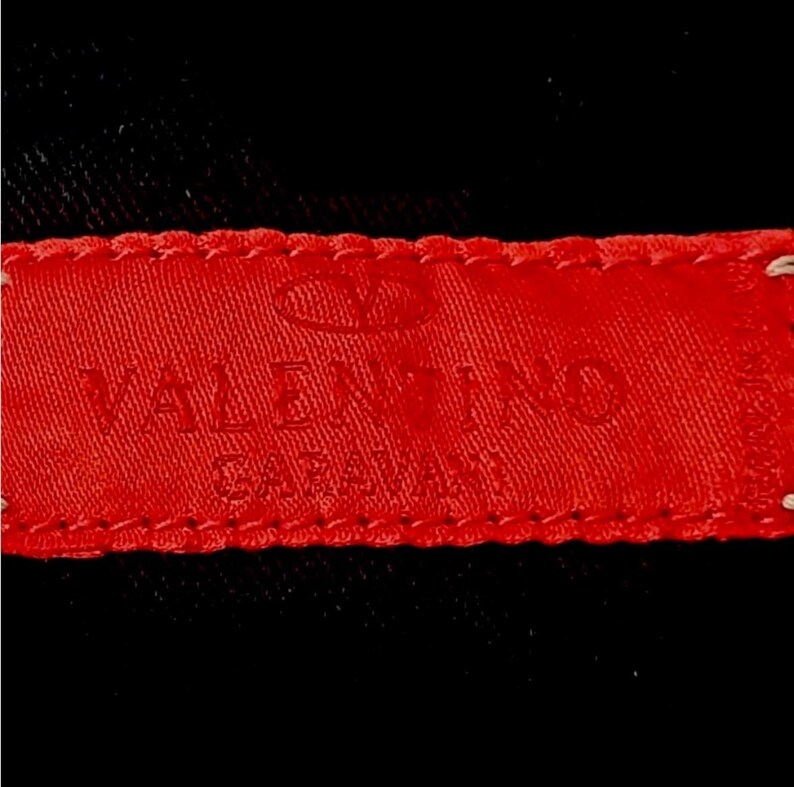 Puede incluir: Primer plano de una etiqueta de tela roja con el texto en relieve "VALENTINO GARAVANI" en may&uacute;sculas. La etiqueta est&aacute; cosida a una tela negra. El logotipo de Valentino es visible en la parte superior de la etiqueta.