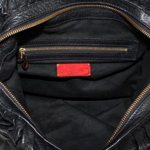 Puede incluir: Bolso de mano de cuero negro con cremallera dorada y etiqueta roja con la inscripci&oacute;n "VALENTINO GARAVANI". El interior es negro con un peque&ntilde;o bolsillo con cremallera. El bolso tiene un dise&ntilde;o texturizado y fruncido.