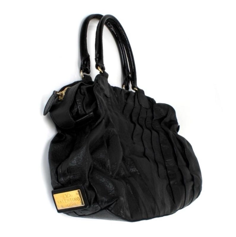 Puede incluir: Bolso de mano de cuero negro con dos asas curvas y una cremallera dorada. El bolso presenta una superficie texturizada y un dise&ntilde;o plisado decorativo en la parte delantera. Una placa rectangular dorada con el nombre de la marca es visible.