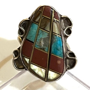 May include: A silver ring with a large, multi-colored stone set in the center. The stone is made of turquoise, red, and white stones, and is set in a geometric pattern. The ring is a statement piece and would be a great addition to any jewelry collection.
