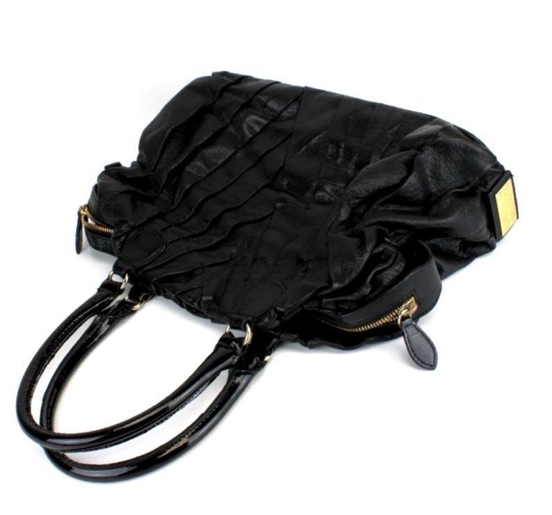 Puede incluir: Bolso de mano de cuero negro con un dise&ntilde;o plisado texturizado. Cuenta con dos asas negras brillantes y cremalleras doradas. Un acento rectangular dorado est&aacute; en el lateral. La bolsa est&aacute; sobre un fondo blanco.