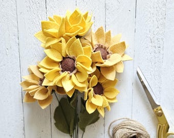 May include: A bouquet of handcrafted felt sunflowers featuring bright yellow petals and textured brown centers. The flowers are attached to thin green stems with dark green felt leaves. Beside the flowers sits a ball of jute twine and gold scissors.