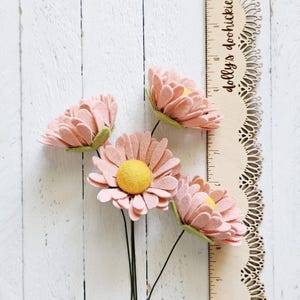 May include: Four handmade felt daisy flowers in shades of pink with yellow centers and green accents. The flowers are on dark green stems. A wooden ruler with the text "dolly's doohickies" is next to the flowers.