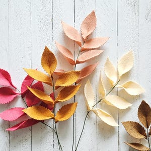 May include: A collection of felt leaf stems in various colors, including pink, yellow, peach, cream, and brown. The leaves are arranged on a white wooden surface, creating a natural and decorative display. These artificial leaves are ideal for crafting and home decor.