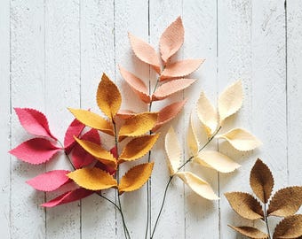 May include: A collection of felt leaf stems in various colors, including pink, yellow, peach, cream, and brown. The leaves are arranged on a white wooden surface, creating a natural and decorative display. These artificial leaves are ideal for crafting and home decor.