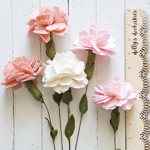May include: Five felt carnation flowers in shades of pink and white with green stems and leaves. The flowers are arranged against a white wooden background. A wooden ruler with the text "dolly's doohickies" is on the right.