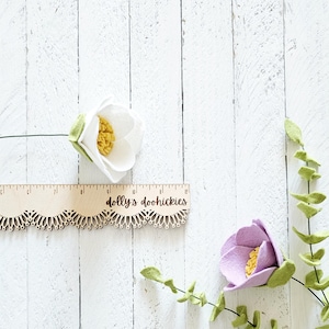May include: Two felt flowers, one white and one purple, with green felt leaves, on a white wooden background. A wooden ruler with the text "dolly's doohickeys" is also in the image.