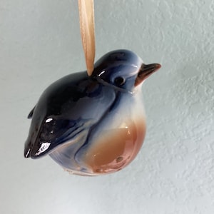 May include: A glossy ceramic bird ornament featuring a dark blue head and back, a white chest, and a light brown belly. The bird has a small dark beak and a black eye, and it is suspended by a thin, light-colored ribbon attached to its back.