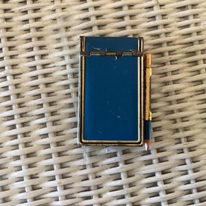 May include: A rectangular blue metal notepad holder with a gold-toned trim and a matching small pencil attached to the side. The notepad holder features a hinged top section and shows signs of wear on the metallic edges and the pencil casing.