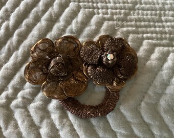 May include: A decorative hair accessory featuring two brown flower-shaped ornaments connected by a ring. The flowers have a textured, woven appearance, with a sparkling gem at the center of one. The accessory is designed for hair styling.