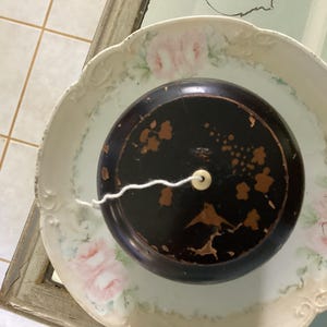 May include: A black, round antique container with gold accents and a white string, resting on a decorative, scalloped-edge plate with pink rose patterns. The plate is white with a cream-colored border.