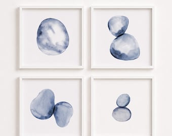 May include: A set of four square white frames containing watercolor paintings of smooth, rounded stones in varying shades of indigo blue. Each frame displays a different composition of one or two abstract, organic pebble shapes on a white background.