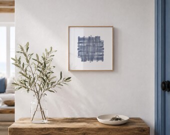 May include: A framed abstract art print with a blue watercolor design hangs on a white wall. The artwork features a woven pattern in shades of blue. A wooden table holds a glass vase with greenery and a small white dish.
