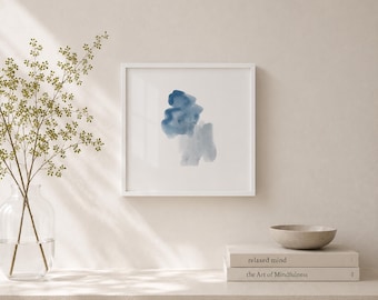 May include: A square white frame containing an abstract watercolor painting with blue and grey shapes. Below the frame are two stacked books titled "relaxed mind" and "the Art of Mindfulness" next to a small grey bowl and a clear glass vase.