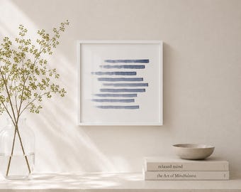 May include: A square white-framed wall art piece featuring horizontal blue watercolor-style brushstrokes. Below the art, a stack of two books titled "relaxed mind" and "the Art of Mindfulness" sits next to a small, shallow beige bowl.
