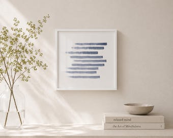 May include: A framed square art print featuring a series of horizontal blue brushstrokes. The artwork is displayed on a white wall, with a vase of greenery and a small bowl on a shelf below. The books "relaxed mind" and "the Art of Mindfulness" are also on the shelf.
