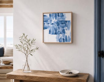 May include: A framed abstract watercolor painting with shades of blue on a white background hangs on a white wall. The painting is in a light brown wooden frame. A wooden bench holds a clear glass vase with greenery and a small white bowl.