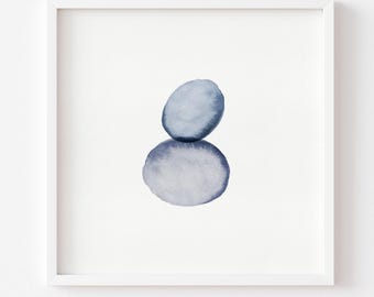 May include: A square white frame containing a watercolor painting of two stacked smooth stones. The top stone is a smaller, rounded blue shape, while the bottom stone is a larger, oval-shaped blue form. The artwork features soft blue gradients.