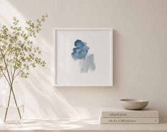 May include: A framed abstract watercolor painting with shades of blue. The artwork is in a white frame and rests on a white surface. A vase with greenery and two books are also on the surface. The books are titled "relaxed mind" and "the Art of Mindfulness".