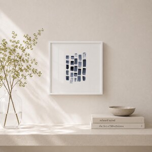 May include: A square white frame containing an abstract painting of dark blue vertical brushstrokes arranged in a grid-like pattern. The artwork is displayed alongside a clear glass vase with green stems and a stack of two books with white covers.