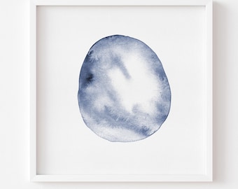 May include: A square white frame containing a watercolor painting of a single, irregular circular shape. The shape features varying shades of indigo and slate blue, with soft, blended edges and lighter white areas within the pigment.