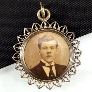 Op de afbeelding: Antieke goudkleurige hanger met een cirkelvormig portret van een man in pak en stropdas. Het portret is omgeven door een decoratief, zonnestraalvormig frame. De hanger heeft een lus voor een ketting.