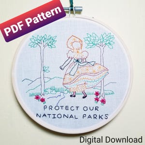 May include: Embroidered hoop art featuring a girl in a yellow dress and a green hat, standing in a forest with the text "Protect Our National Parks".