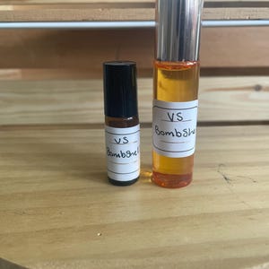 May include: Two perfume bottles of different sizes. The larger bottle has a silver cap and contains a golden liquid. Both bottles have a white label with the handwritten text "VS Bombshell". The bottles are on a wooden surface.