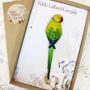 May include: A green and yellow embroidered bird brooch with a red beak, on a white background with floral designs. The card reads "Vikki Lafford Garside".