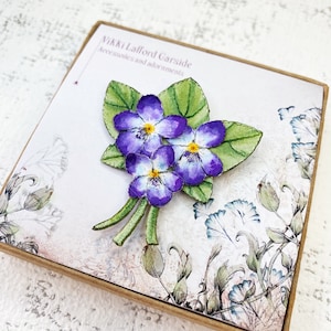 May include: A fabric brooch depicting three purple violets with yellow centres and green leaves. The brooch is displayed on a card with the text "Nikki Lafford Garside Accessories and adornments".