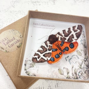 May include: A brown and white moth with orange wings and blue spots is displayed in a small box. The box is lined with white paper and has a floral pattern. The box is labeled "Vikki Lafford Garside, Accessories and adornments."