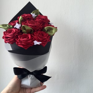 May include: A bouquet of red satin roses with green leaves and white accents wrapped in black paper and tied with a black ribbon.