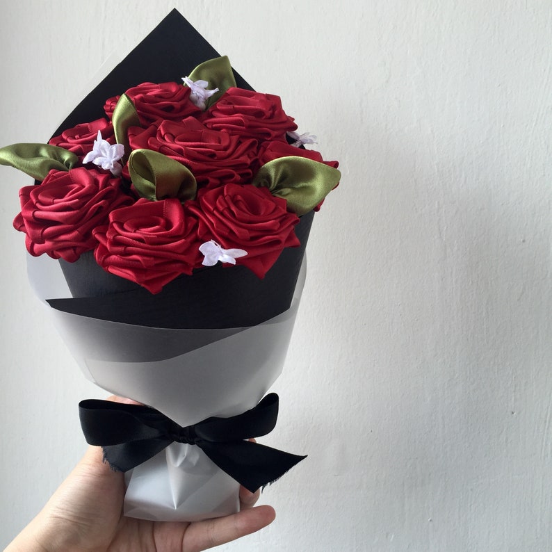 May include: A bouquet of red satin roses with green leaves and white accents wrapped in black paper and tied with a black ribbon.