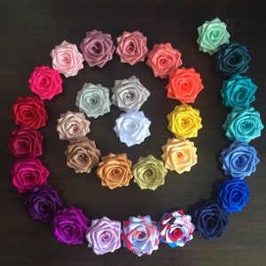 May include: A collection of 25 handmade satin fabric rose flower embellishments in various colors, including pink, red, orange, yellow, green, blue, purple, and white. The roses are arranged in a spiral pattern.
