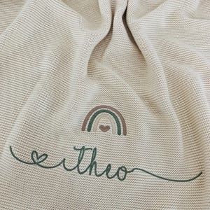 May include: A beige knitted blanket featuring an embroidered design of a small rainbow with a heart at its center. Below the rainbow, the name "theo" is stitched in dark green cursive script, preceded by a small heart outline.