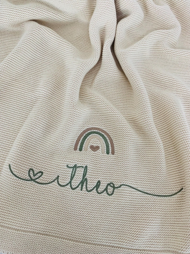 May include: A beige knitted blanket featuring an embroidered design of a small rainbow with a heart at its center. Below the rainbow, the name "theo" is stitched in dark green cursive script, preceded by a small heart outline.