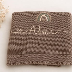 May include: A brown knitted blanket featuring an embroidered rainbow design at the top center and the name Alma stitched in cursive script across the middle, accompanied by a small heart icon to the left of the name in a light cream thread.