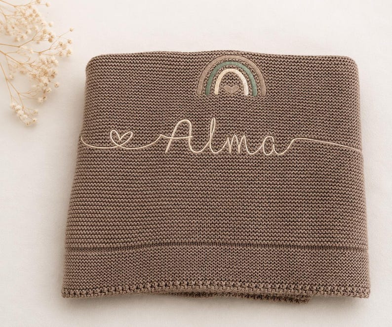 May include: A brown knitted blanket featuring an embroidered rainbow design at the top center and the name Alma stitched in cursive script across the middle, accompanied by a small heart icon to the left of the name in a light cream thread.