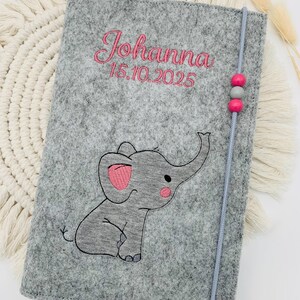 May include: A gray felt notebook cover featuring an embroidered elephant with pink accents. The name Johanna and the date 15.10.2025 are stitched in pink thread. A gray elastic cord with two pink beads and one gray bead secures the cover.