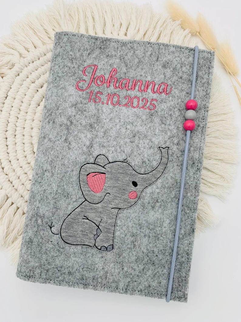 May include: A gray felt notebook cover featuring an embroidered elephant with pink accents. The name Johanna and the date 15.10.2025 are stitched in pink thread. A gray elastic cord with two pink beads and one gray bead secures the cover.