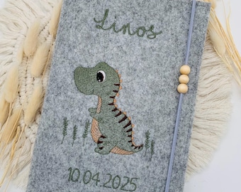 May include: A gray felt notebook cover featuring an embroidered green dinosaur, the name Linos, and the date 10.04.2025. The cover includes a light blue elastic closure band adorned with three small, round, light-colored wooden beads.