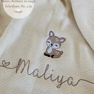 May include: A natural-colored knit blanket featuring an embroidered taupe fawn graphic and the name Maliya in cursive taupe script. Text on the blanket reads: Deckenfarbe: Natur, Motiv: Rehkitz in taupe, Schriftart: Nr. 2 in taupe.